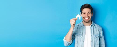 Real estate concept. Young handsome man smiling, showing small paper house mockup, searching for apartment, standing over blue background photo