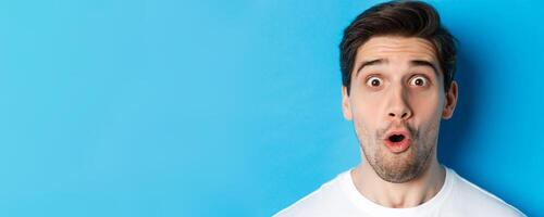 Headshot of surprised man gasping wondered, saying wow and looking amazed at camera, standing over blue background photo
