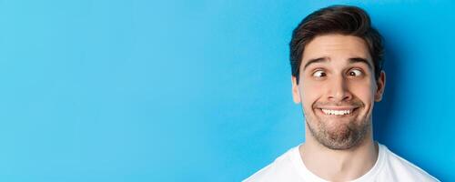 Head shot of young man making funny expressions, smiling and squinting, standing over blue background photo