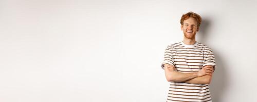 Happy redhead guy with bristle laughing and smiling at camera, standing with hands crossed on chest over white background photo