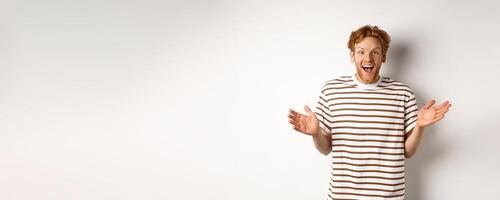 Happy and surprised redhead man rejoicing of good news, looking amazed and pleased at camera, white background photo