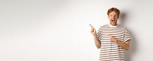Cheerful man with red hair checking out promo offfer, pointing left at logo and smiling at camera, standing over white background photo