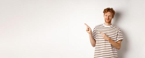 Doubtful redhead guy pointing fingers left at looking skeptical at camera, standing over white background photo