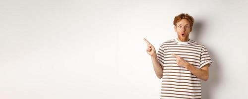 Amazed man with ginger hair checking out promo offfer, pointing left at logo and smiling at camera, standing over white background photo