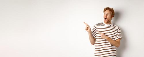 Check it out. Amazed redhead guy pointing and looking left at promotion banner, standing over white background photo