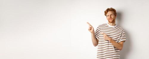 Doubtful young man with bristle and red hair looking, pointing fingers left at something average, staring skeptical at promo, white background photo