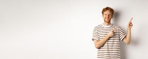 Handsome redhead man showing logo, pointing fingers right and smiling, standing over white background photo