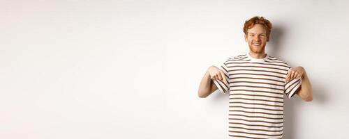 Smiling young man with messy red hair and beard, pointing fingers down, recommending shop, standing over white background photo