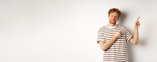 Judgemental redhead guy frowning disappointed, pointing fingers upper right corner at something bad, white background photo