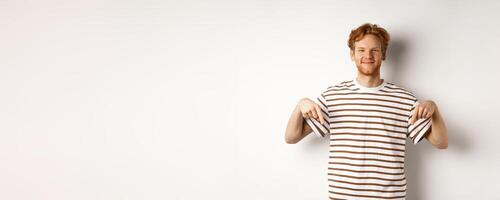 Handsome male model with ginger hair and bristle, pointing fingers down and smiling, showing advertisement, white background photo