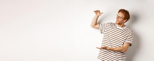 Happy and satisfied caucasian redhead man introduce something big, showing large product and smiling pleased, white background photo