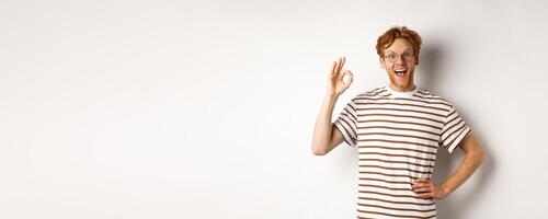 Excellent job. Happy redhead man praising you, showing OK sign and smiling amazed, approve and like something good, standing over white background photo