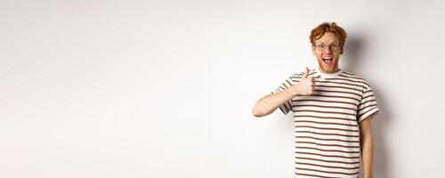 Amazed and fascinated redhead guy checking out something awesome, showing thumb-up and scream yes with joy, standing over white background photo