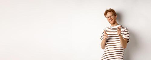 Cool and sassy redhead man with beard and glasses, pointing finger pistols at camera and smiling, congratulating you, standing over white background photo