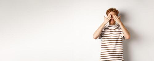 Smiling man cover eyes and waiting for surprise, standing in t-shirt against white background photo
