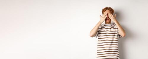 Smiling man cover eyes and waiting for surprise, standing in t-shirt against white background photo