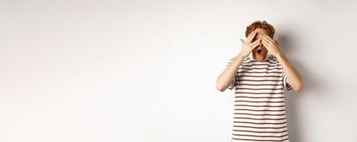 Shocked and embarrassed redhead guy covering eyes, peeking through fingers and gasping, standing over white background photo