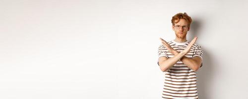 Serious young man with ginger hair frowning, showing stop gesture, making cross to prohibit something bad, disagree with you, standing over white background photo