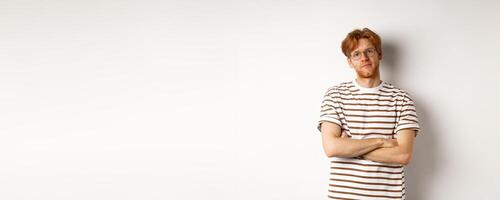 Teenage redhead guy in glasses cross arms on chest, looking skeptical and unamused at camera, standing over white background photo