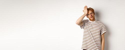 Tired and distressed redhead male student slap forehead, making facepalm and staring at camera exhausted, standing over white background photo
