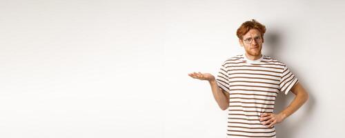 So what. Confused and annoyed redhead man waiting for reply, standing questioned with raised hand, holding something on palm, white background photo