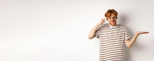 Are you stupid. Confused redhead guy in glasses shrugging, pointing at head and staring skeptical at camera, white background photo