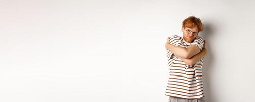 Reluctant young man with messy red hair unwilling do something, hugging himself and grimacing, standing over white background photo
