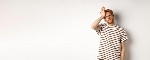 Tired young man with red hair and glasses, roll eyes and making facepalm bothered, standing over white background photo