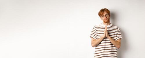 Silly redhead man asking for help, holding hands in pleading gesture and pucker lips, say please while begging you, standing over white background photo