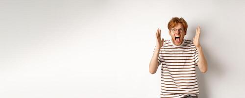 Angry young man with red hair shouting at camera, screaming and looking outraged, shaking hands, standing over white background photo
