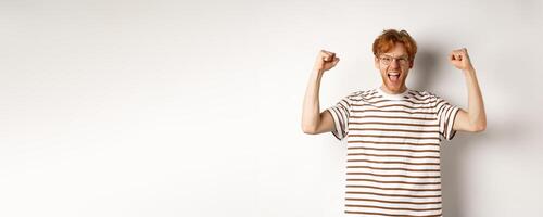 Joyful redhead guy raising hands up like winner, winning prize and celebrating, shouting for joy, standing over white background photo
