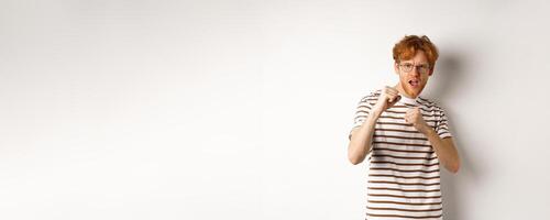 Funny young man with red hair raising fists for fight, shadow boxing and looking serious at camera, standing over white background photo
