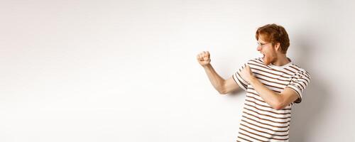 Profile of funny young man with red hair and beard, raising fists for fight, shadow boxing and shouting, standing over white background photo
