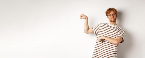 Image of confident and strong redhead man flexing biceps, showing muscles after gym, standing over white background photo