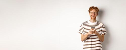 Technology and e-commerce concept. Redhead guy in glasses using mobile phone and smiling. Young man with smartphone staring happy at camera, white background photo