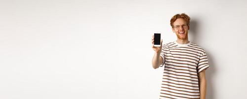 Technology and e-commerce concept. Happy young redhead man in glasses showing blank smartphone screen, smiling satisfied, standing over white background photo