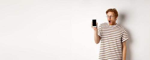 Technology and e-commerce concept. Surprised and shocked redhead guy checking out online promotion, showing blank smartphone screen and drop jaw, white background photo