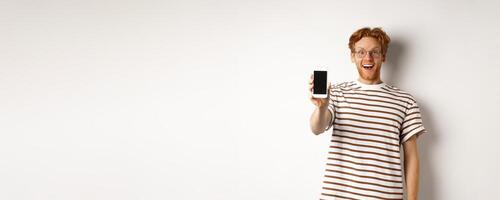 Technology and e-commerce concept. Happy young redhead man in glasses showing blank smartphone screen, looking at camera amazed, standing over white background photo