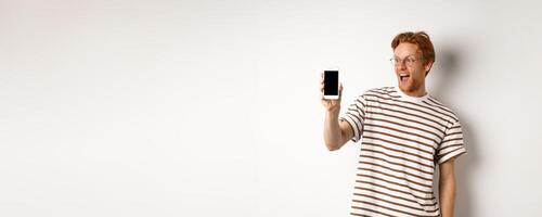 Technology and e-commerce concept. Happy young redhead man in glasses showing blank smartphone screen, looking amazed, standing over white background photo