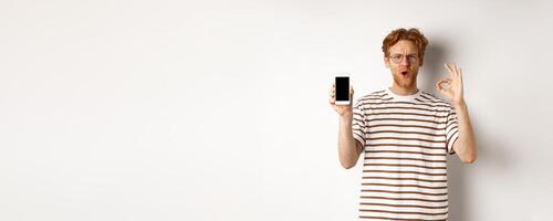 Technology and e-commerce concept. Young man with red hair showing okay sign and blank smartphone screen, praising awesome app or game, white background photo