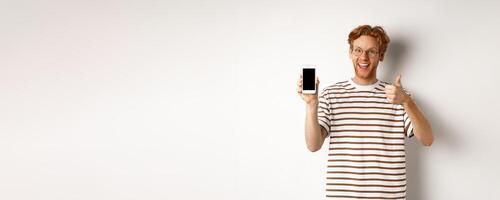 Technology and e-commerce concept. Young man with red hair showing thumbs-up and blank smartphone screen, recommending app, white background photo