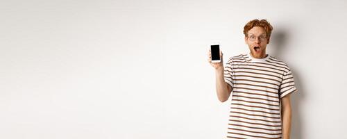 Technology and e-commerce concept. Surprised and shocked redhead guy checking out online promotion, showing blank smartphone screen and drop jaw, white background photo