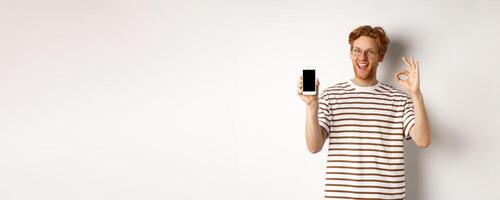 Technology and e-commerce concept. Young man with red hair showing okay sign and blank smartphone screen, praising awesome app, standing over white background photo