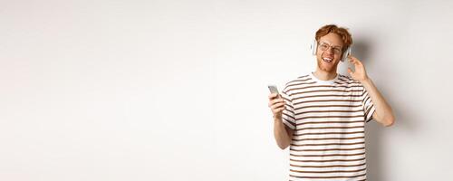 Technology concept. Young man with red hair and beard listening music in headphones and using smartphone, smiling at camera, white background photo