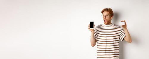 Shopping and finance concept. Amazed young man with red hair showing plastic credit card and smartphone blank screen, staring at display impressed, white background photo