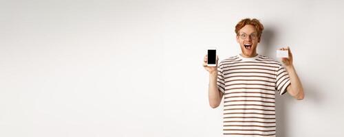 Shopping and finance concept. Young man showing blank mobile screen and plastic credit card, smiling at camera, white background photo