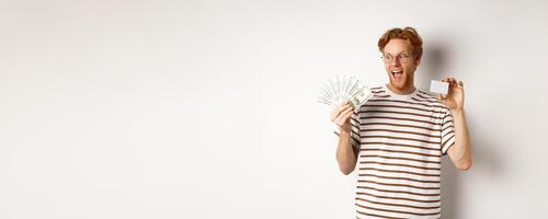 Shopping and finance concept. Cheerful handsome redhead man showing plastic credit card and cash, smiling amazed, white background photo