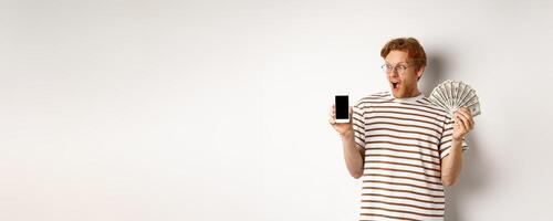 Amazed redhead man showing smartphone app on blank screen and money, winning prize cash online, standing over white background photo