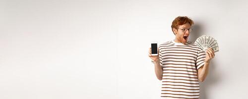 Amazed redhead man showing smartphone app on blank screen and money, winning prize cash online, standing over white background photo