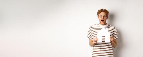 Happy and surprised redhead man winning house, holding paper home model and staring at camera, standing joyful over white background photo